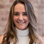 A woman with long, wavy hair wears a white sleeveless top in front of a softly blurred brick background.