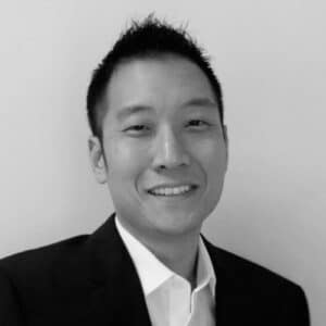 A man in a black suit and white shirt poses against a plain background, exuding a professional appearance.