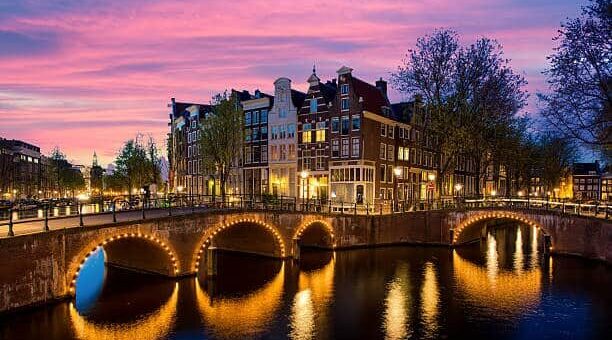Twilight over Amsterdam canal with illuminated bridge and historic buildings.