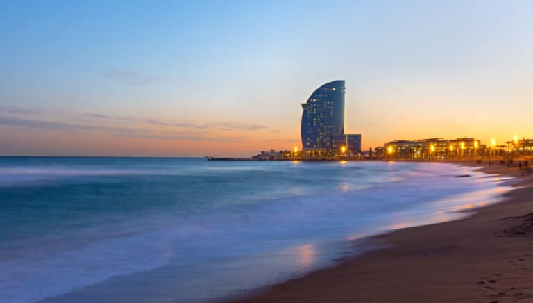 Twilight beach scene with illuminated skyscraper and smooth water reflections.