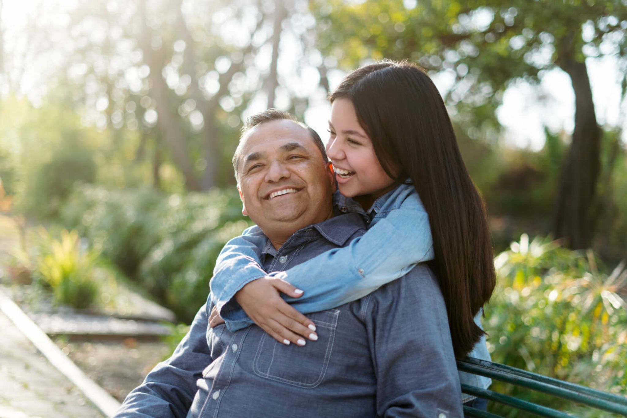 A daughter hugging her father who is sitting on a bench and smiling up to her