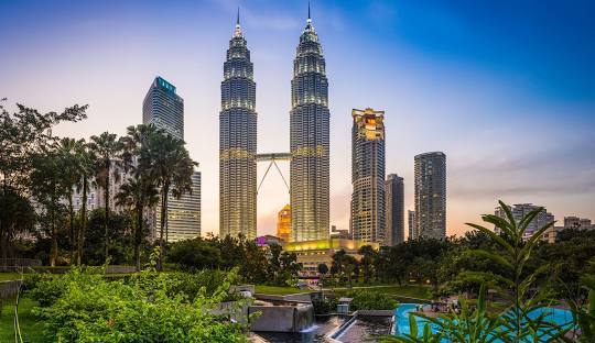 The iconic Petronas Towers rise majestically at sunset, surrounded by lush greenery and modern cityscape in Kuala Lumpur.