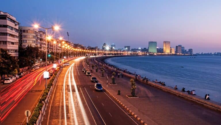 A scenic view of a city waterfront promenade at twilight, featuring bustling traffic and people strolling along the shore.