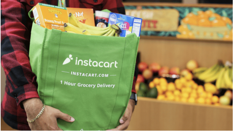 Person holding a green Instacart grocery delivery bag filled with various food items, with fruit stands in the background.
